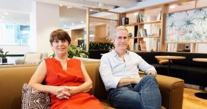 A woman in a red dress sits next to a man in a button-up shirt and jeans on a couch in a co-working space in Brisbane's CBD