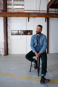 A Southeast Asian man smiling, sitting on a stool in a warehouse-style room with a piece of modern art on the wall behind him