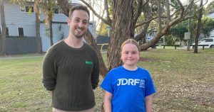 Stephen Bates stands next to Sophie, a young girl who is wearing a blue 'JDRF' shirt, in a park